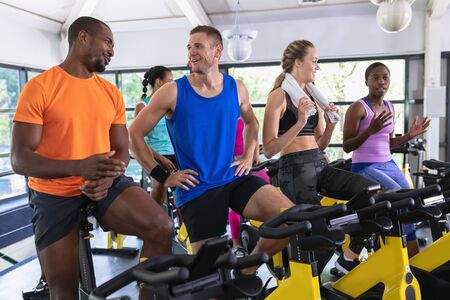 Front view of diverse fit people interacting with each other exercising on exercise bike in fitness center. Bright modern gym with fit healthy people working out and training at spin classの写真素材