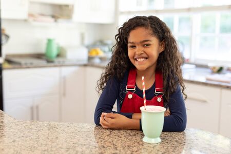 Portrait of a young mixed race girl sitting in a kitchen at Christmas, smiling to camera with a mug...