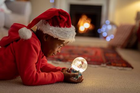 Side view of a young mixed race boy in his sitting room at Christmas, wearing a santa hat and holding a snow globeの写真素材