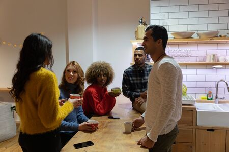 Side view of a group of young adult multi-ethnic male and female friends socialising in the kitchen of an apartment, talking together and drinking coffeeの写真素材