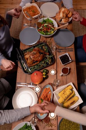 High angle view of a group of young adult multi-ethnic male and female friends sitting around a table holding hands saying grace before eating Thanksgiving dinner at home togetherの写真素材