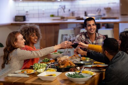 Side view of a group of young adult multi-ethnic male and female friends sitting at a table at home set for Thanksgiving dinner making a toast with glasses of red wineの写真素材