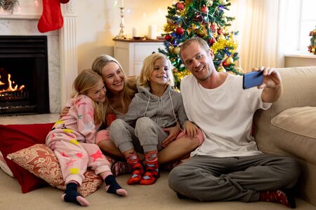 Front view of a Caucasian couple sitting on the floor with their young son and daughter in their sitting room at Christmas time, dad taking a selfie with a smartphone, a decorated Christmas tree in the backgroundの写真素材