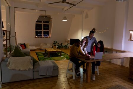 Front view of a group of four young multi-ethnic male and female friends looking at a laptop computer together, talking and laughing in the sitting room of an apartment at nightの写真素材