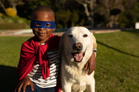 Portrait of a young african american boy dressed in a superhero costume with a red cape and a blue mask in a garden, embracing a dog and smiling to cameraの写真素材