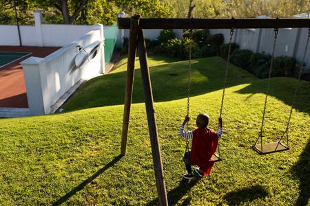 Rear view of a young african american boy dressed in a superhero costume with a red cape and a blue mask in a garden, sitting on a swingの写真素材