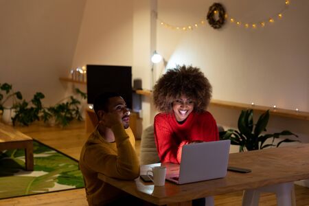 Front view of a young mixed race man and woman sitting at a table looking at a laptop computer together and smiling in the sitting room of an apartmentの写真素材