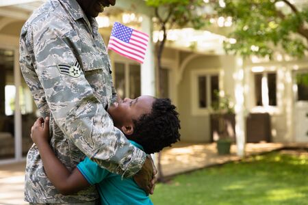 Side view close up of a young adult African American male soldier in the garden outside his home, embracing his young son, who is looking up at him smiling and holding a US flag, thier house in the backgroundの写真素材