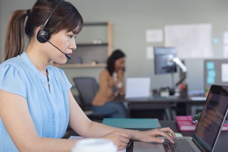 Side view close up of an Asian female business creative working in a casual modern office, sitting at a desk using a laptop computer and wearing a phone headset, with colleagues working in the backgroundの写真素材