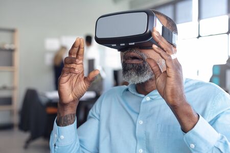 Portrait of an African American male business creative working in a casual modern office, sitting and using a VR headset and raising his hands up in front of him, with colleagues in the backgroundの写真素材