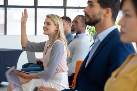 Side view of a diverse group of business creatives sitting in a row in the audience at a business conference, with one blonde haired Caucasian young woman smiling and raising her hand to ask a questionの写真素材