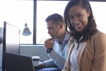 Portrait of a mixed race female business creative working in a casual modern office smiling to camera, with a Caucasian male colleague sitting at a desk working with a computer in the backgroundの写真素材