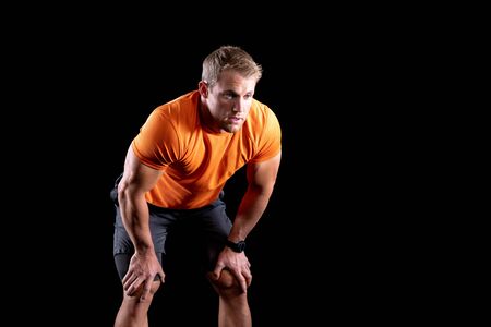 Front view of a muscular Caucasian man wearing sports clothes recovering after a work out, leaning forward with his hands on his knees. On a black backgroundの写真素材