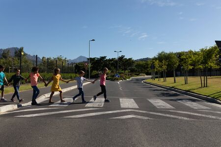 Side view of a diverse group of elementary school pupils crossing an empty road together, half way across a pedestrian crossing, holding hands on a sunny dayの写真素材