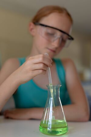 Front view close up of a Caucasian elementary school girl sitting at a table wearing safety glasses and using a pipette and a beaker to do an experiment during a chemistry lesson, focus on the foregroundの写真素材