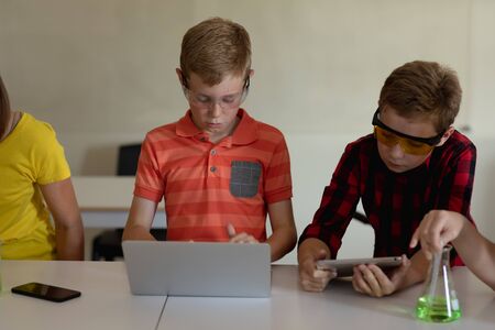Front view of two Caucasian schoolboys sitting at a desk wearing safety glasses and using a tablet and a laptop computer during an elementary school science class, with classmates working beside themの写真素材