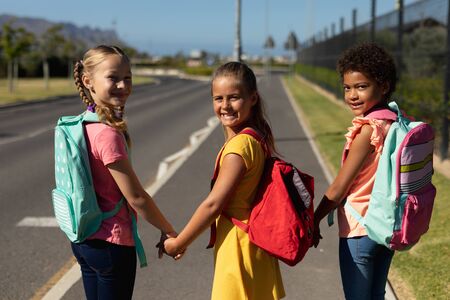 Rear view of two Caucasian schoolgirls and an African American schoolgirl walking along a road to...