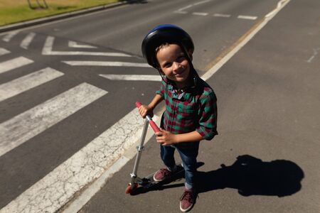 Portrait of a Caucasian schoolboy wearing a helmet and standing with a scooter smiling to camera while waiting at a pedestrian crossing to cross the road safely on his way to elementary school on a sunny dayの写真素材