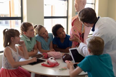 Rear view of a Caucasian male teacher wearing a lab coat using a human anatomy model to teach a diverse group of elementary school children about human organs during a biology lesson, the children sitting in a circle at a table and listening while he talks, holding a model organの写真素材
