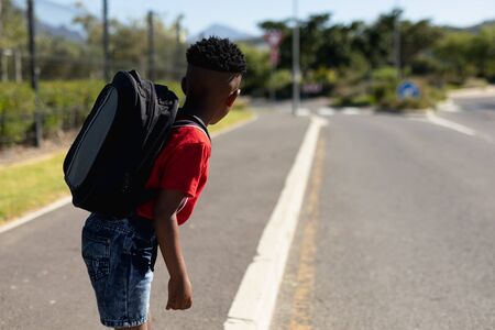 Side view of an African American schoolboy wearing shorts and a t shirt and carrying a rucksack, looking away from camera for traffic while waiting to cross the road on the way to elementary school on a sunny dayの写真素材