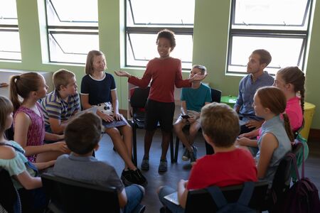 Front view of a diverse group of elementary school kids sitting on chairs in a circle and interacting during a lesson, one African American girl standing and talking while her classmates and male Caucasian teacher sit and listenの写真素材