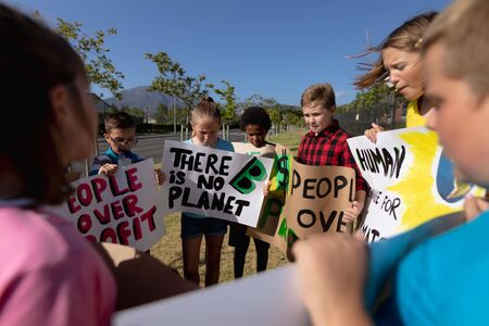 Over the shoulder view of a diverse group of elementary school pupils on a protest march, carrying signs with environmental and conservation slogans on them, walking down a road in the sunの写真素材