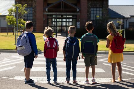 Rear view of a group of three Caucasian schoolboys and a schoolgirl carrying rucksacks waiting at a pedestrian crossing to cross the road in front of their elementary school on a sunny dayの写真素材