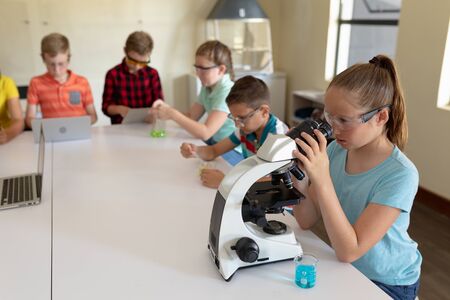 Side view of a Caucasian elementary school girl wearing safety glasses and a blue t shirt, sitting at a desk using a microscope during a science class, with classmates working together sitting in the backgroundの写真素材