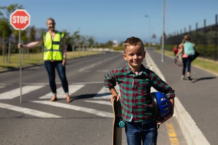 Portrait of a Caucasian schoolboy standing by the road holding a skateboard and a helmet and smiling to camera, a Caucasian woman wearing a high visibility vest and holding a stop sign standing in the road on a pedestrian crossing, stopping the traffic in the backgroundの写真素材