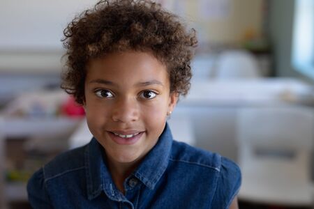 Portrait close up of an African American schoolgirl with short curly hair wearing a blue denim dress, looking to camera smiling in an elementary school classroomの写真素材