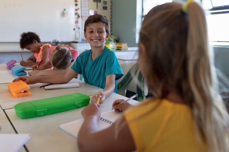 Front view of a Caucasian schoolboy wearing a blue t shirt, sitting at a desk and smiling to camera,...