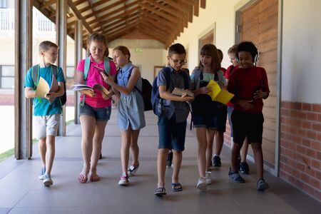 Front view of a diverse group of school pupils wearing rucksacks, carrying books, using phones and...