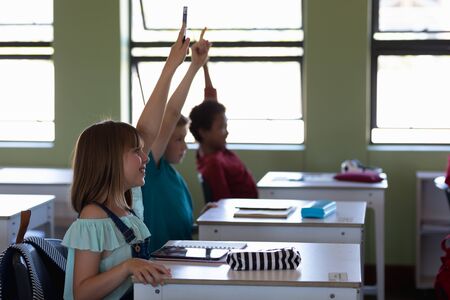 Side view of a diverse group of school children sitting at desks and raising their hands to answer a...