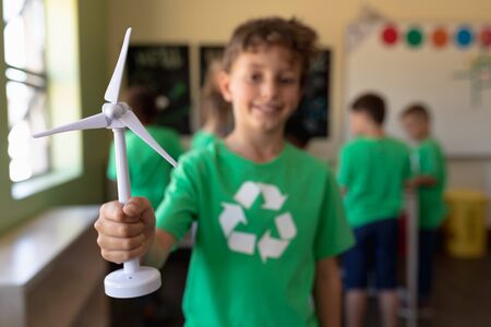Front view of a Caucasian schoolboy with short dark hair wearing a green t shirt with a white recycling logo on it, holding miniature wind turbine and looking to camera smiling in an elementary school classroom, with his classmates standing in the background, focus on the foregroundの写真素材
