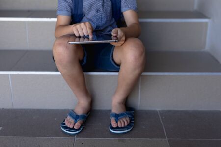 Low section of a Caucasian schoolboy wearing flip-flops and shorts sitting on steps at elementary school using a tablet computerの写真素材