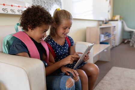 Side view of a Caucasian and an African AMerican schoolgirl sitting on a seat wearing their...