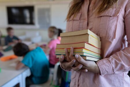 Front view mid section of a Caucasian female elementary school teacher with long blonde hair standing in a classroom holding a pile of books, with her pupils sitting at a table working in the backgroundの写真素材