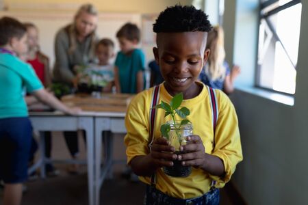 Portrait close up of an African American schoolboy wearing a yellow shirt holding a seedling planted in a jar of earth, looking at it and smiling during a nature study lesson in an elementary school classroom, with his teacher and classmates in the backgroundの写真素材