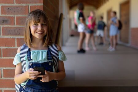 Front view of a Caucasian schoolgirl wearing a rucksack leaning against a wall in the schoolyard at elementary school using a smartphone, looking to camera and smiling, with other schoolchildren standing together in the backgroundの写真素材