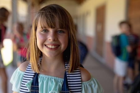 Portrait close up of a Caucasian schoolgirl wearing a rucksack standing in the schoolyard at elementary school smiling to camera, with other schoolchildren standing in the backgroundの写真素材