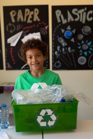 Portrait of an African American schoolgirl with short hair wearing a green t shirt with a white recycling logo on it, holding a recycling crate and looking to camera smiling in an elementary school classroom, with poster about recyclable materials in the backgroundの写真素材