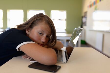 Side view close up of a tired Caucasian schoolgirl sitting at a desk leaning on her laptop computer...