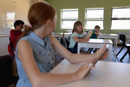 Side view close up of a Caucasian schoolgirl with red hair wearing a blue dress sitting at a desk using a tablet computer and looking at another Caucasian schoolgirl sitting at the next desk in an elementary school classroom, with other schoolchildren sitting at desks working in the backgroundの写真素材