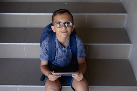 High angle view of a Caucasian schoolboy wearing glasses, shorts and a rucksack sitting on steps at elementary school using a tablet computer, looking up to camera and smilingの写真素材