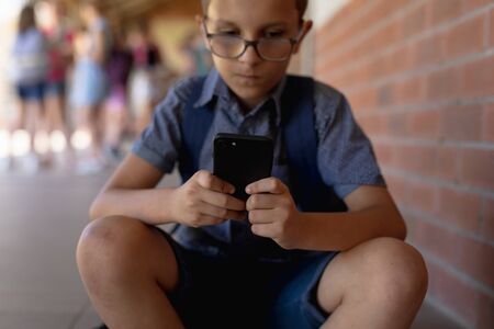 Front view close up of a Caucasian schoolboy wearing glasses, shorts and a rucksack sitting by a...