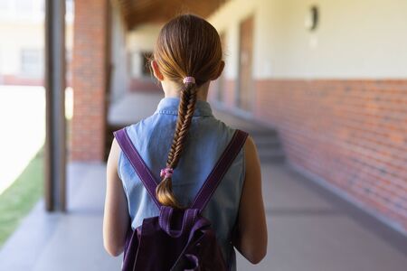 Front view of a Caucasian schoolgirl with red hair in a plait wearing a blue dress and a rucksack...