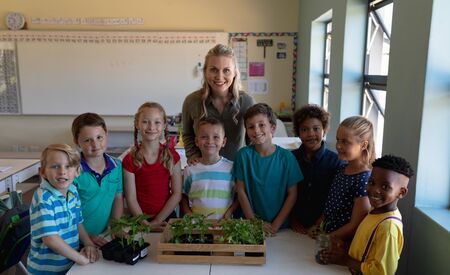 Portrait of a Caucasian female elementary school teacher with long blonde hair and a diverse group of eight schoolchildren gathered around a box of plants for a nature study lesson in an elementary school classroom, smiling to cameraの写真素材
