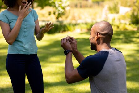 Rear view of an African American man kneeling in the garden and proposing to his mixed race female partner on a sunny dayの写真素材