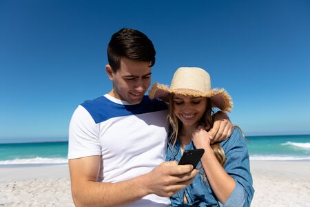 Front view of a Caucasian couple standing on the beach with blue sky and sea in the background, embracing and looking at smartphoneの写真素材