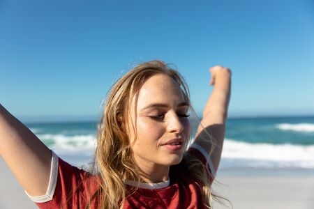 Front view of a Caucasian woman with long blonde hair, standing on the beach with blue sky and sea...